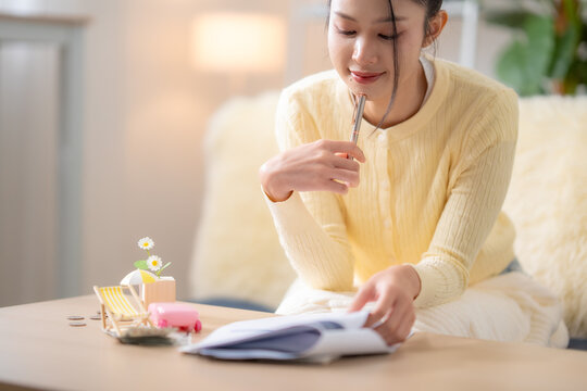 Asian woman reading travel plan beside suitcase, beach chair, coins, saving money for vacation, planning holiday budget, tourism expenses, future trip concept.