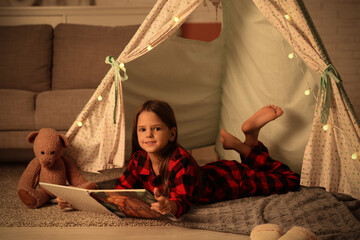 Fototapeta premium Cute little girl reading fairy-tale while lying in play tent at home