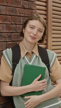 Portrait of young adult Caucasian female student holding book, adjusting hair, and smiling at camera. Clip is ideal for college marketing, vertical