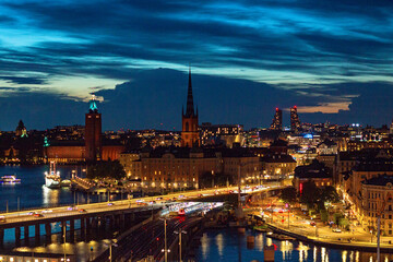 Fototapeta premium Stockholm, Sweden - 09.12.2025: Illuminated Stockholm landmarks viewed from Katarinahissen across Riddarfjärden, featuring City Hall and Riddarholmen Church