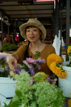 Asian Woman Flower Vendor At Farmers Market Selecting Fresh Blooms In Straw Hat