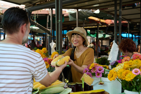 Smiling Asian Woman Vendor Handing Fresh Corn to Customer at Outdoor Farmers Market Stall