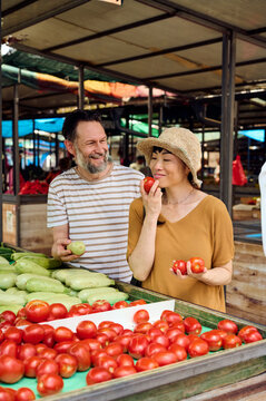 Couple Shopping At Outdoor Farmers Market Smiling While Choosing Fresh Tomatoes And Zucchini