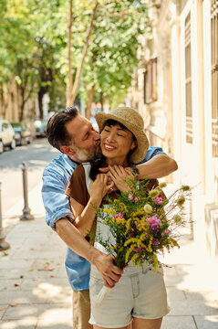 Couple Embracing on Sunny City Street &mdash; Man Kissing Asian Woman Holding Wildflower Bouquet