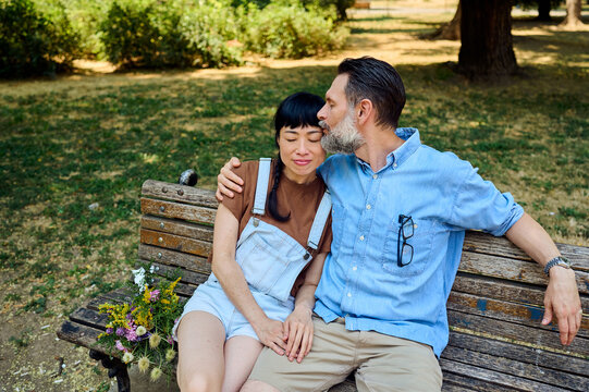 Affectionate Couple Sitting on Park Bench With Flowers, Man Kissing Woman&rsquo;s Cheek on Summer Date