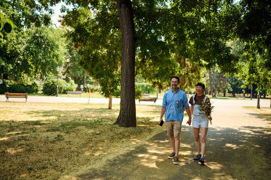 Happy Couple Walking Hand in Hand in Sunny Park With Flowers and Casual Summer Style