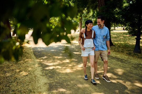 Happy Couple Walking Arm in Arm Through Sunny City Park &mdash; Asian Woman and Partner on Summer Stroll