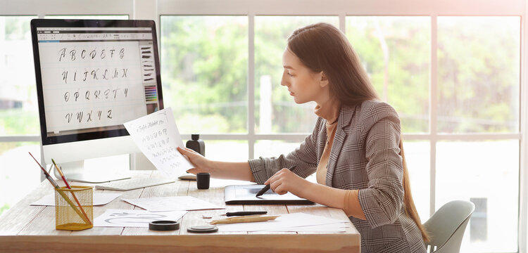 Young female graphic designer creating custom calligraphy font on computer with drawing tablet in bright studio, She holds a reference sheet of handwritten letters