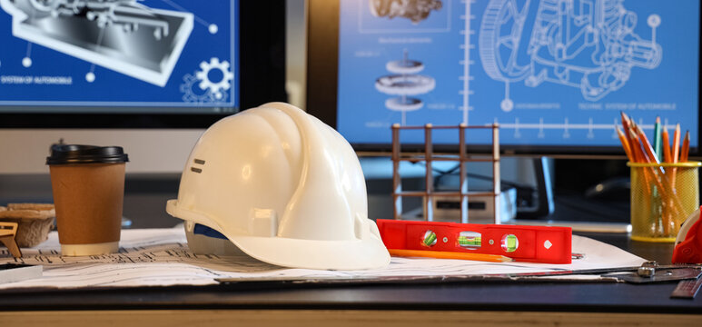 White hard hat and blueprints on an engineer's desk with computer screens displaying technical drawings and design plans