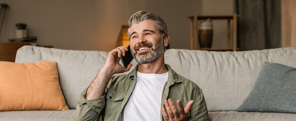 Fototapeta premium Happy Senior Man Smiling While Talking On Smartphone In Modern Living Room, Engaged In Conversation At Home. Cheerful Mature Caucasian Male Enjoying Interaction In Comfortable Setting.