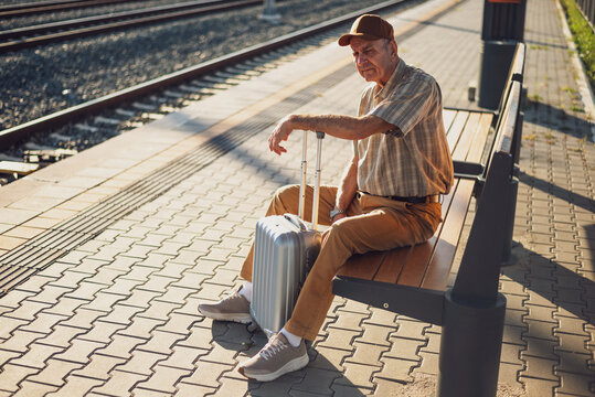 Tired and bored senior man is sitting on bench at railway station and waiting for arrival of the train. Senior people traveling.
