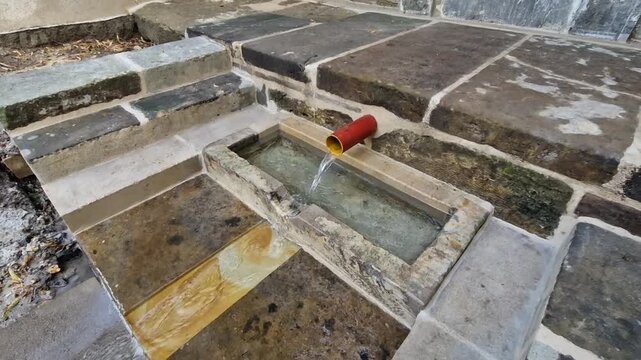 natural spring or well area, featuring a small stone trough where water streams out of a red outlet pipe. The surrounding structure consists of large, rustic stone slabs forming steps and paving aroun