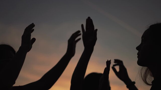 Hands raised in silhouette at sunset. Group of people wave arms against dusk sky. Friends raise hands together in celebration. Silhouette of hands moving at twilight. People enjoy sunset gathering.