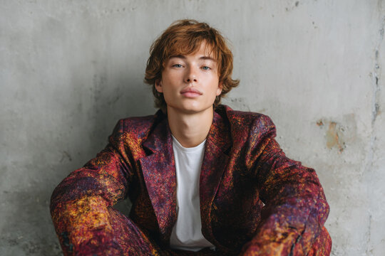 Young man with auburn hair wearing a bold red abstract print blazer against raw concrete wall