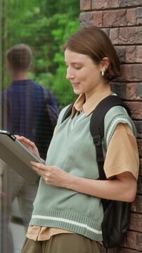 Portrait of Caucasian female teenager leaning against wall, using tablet and smiling on college campus. Ideal for promoting educational apps or university enrollment campaigns, vertical