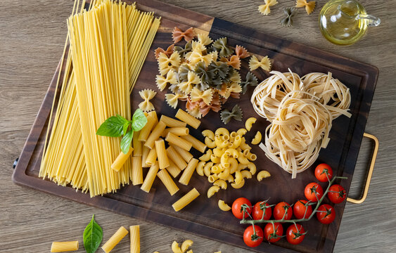 Assorted Italian pasta varieties arranged on a rustic wooden cutting board, including spaghetti, penne, macaroni, farfalle, tagliatelle, fresh basil, cherry tomatoes, red onion, olive oil,