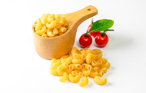 Uncooked macaroni pasta arranged in a wooden scoop with loose pasta pieces, fresh cherry tomatoes, and basil leaves on a clean white background. 