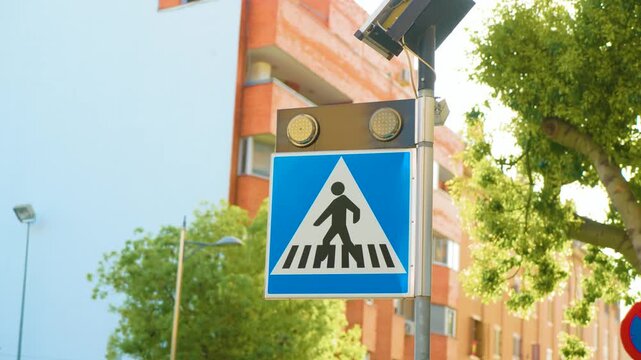 Solar powered pedestrian crossing sign beside residential buildings in Spain. LED warning lights above blue crosswalk symbol. Urban road safety infrastructure on sunny street