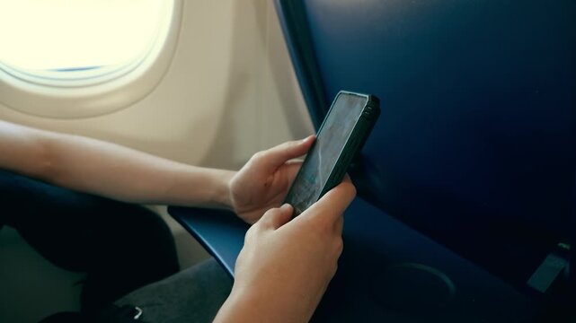 Unrecognizable passenger typing text messages on smartphone beside airplane window. Hands using mobile phone during flight. Digital communication and travel lifestyle concept