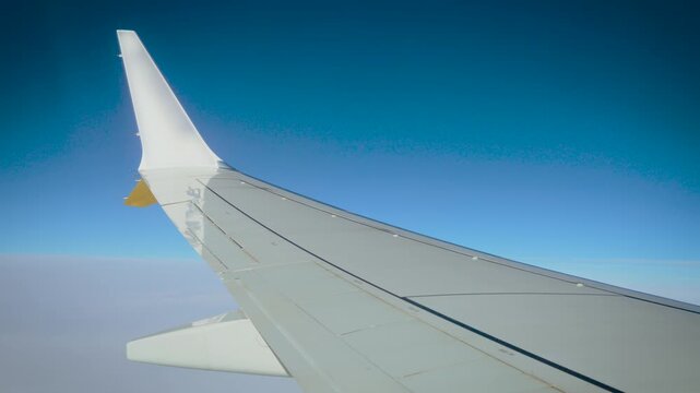 Aircraft wing view through airplane window over soft cloud layer. Bright blue sky above white cumulus clouds at high altitude. Passenger flight perspective during air travel