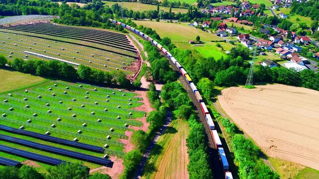 Aerial view of a colourful container freight train passing a large hillside solar farm under construction beside a German village and harvested summer fields.