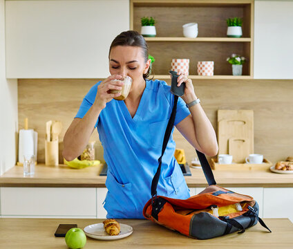 Portrait of a young nurse getting ready for work and using smartphone phone for call online meeting standing in kitchen home. Overworked, busy, stress, urgency, multitasking concepts