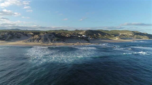 Drone slowly turns and pulls away at Playa El Rinconcito, showing off the dunes with a sliver of landscape becoming visible behind them, and more of the beach as waves roll onto the shore and over a r