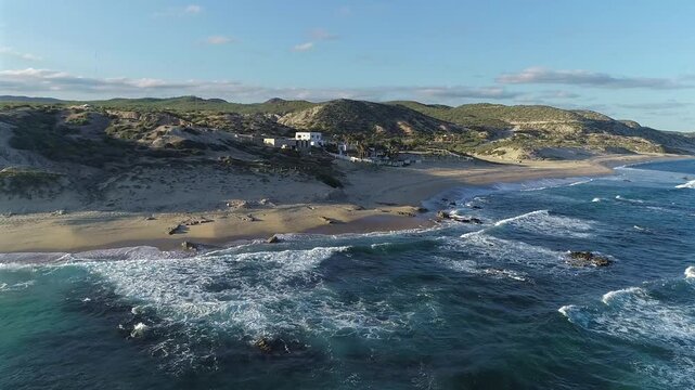 Drone pushes in over the water and rocks at Playa El Rinconcito, approaching a house on the beach and the sand dunes behind it while staying far enough out that the ocean remains visible throughout th