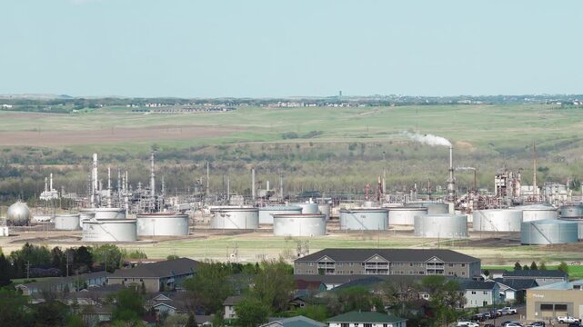 Aerial view of oil storage tanks and silos at a petroleum refinery in Mandan, North Dakota, with a smokestack plume and green prairie hills under a blue sky.