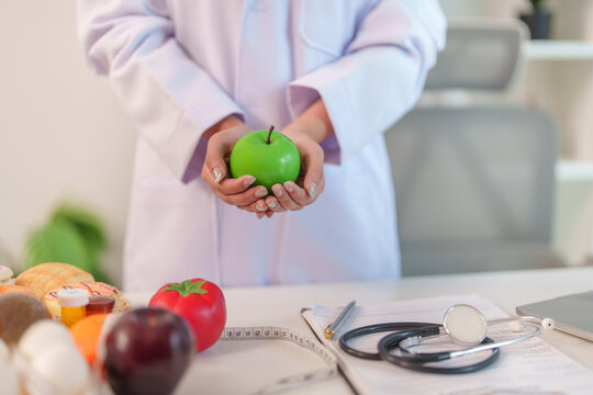 Nutrition consultant holding green apple in clinic with stethoscope on desk