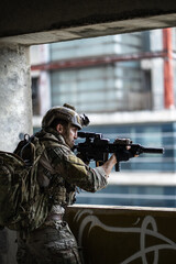 Fototapeta premium Armed tactical soldier aiming rifle over balcony wall wearing camouflage uniform backpack helmet inside concrete building during urban warfare mission