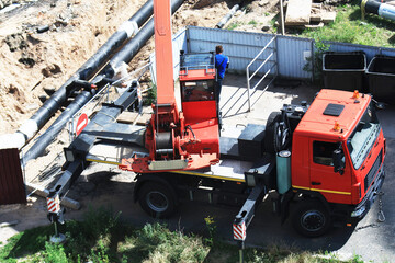 Red truck crane performing work on construction site, heavy machinery with boom and outriggers extended, worker overseeing operation near trench with underground pipes, daytime outdoor scene © HENADZY