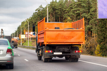 Rear view of orange dump truck driving on multi-lane highway with forest alongside road under cloudy sky © HENADZY