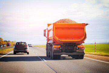 Orange dump truck loaded with gravel driving on highway under bright sky. Construction vehicle transporting materials on asphalt road. Heavy machinery on open route with other cars in background. © HENADZY