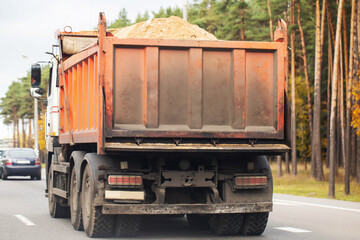 Large orange dump truck transports sand on road, vehicle travels near forest of pine trees and autumn foliage © HENADZY