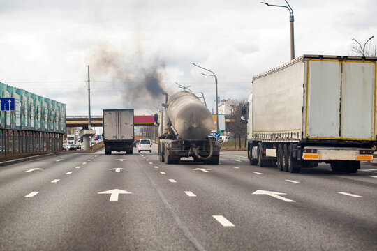 Trucks and cars traveling on multi-lane urban highway under overcast sky, tanker truck emitting black exhaust pollution into air.