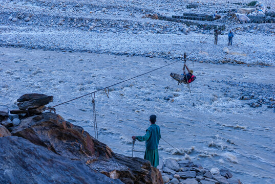 Askole, Gilgit Baltistan, Pakistan - 10 June 2007: View of a person crossing the Braldu River in a jhoola basket on a cable, with a man operating the rope on the rocky riverbank.