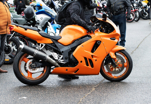 Orange sport motorcycle parked on wet asphalt, rider in black gear and helmet stands beside vehicle, blurred background shows many other bikes and people gathered at outdoor event.