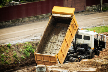 Heavy construction dump truck with yellow cargo bed elevated on muddy site, indicating material unloading or readiness for transport operation, outdoor scene of industrial work, copy space © HENADZY