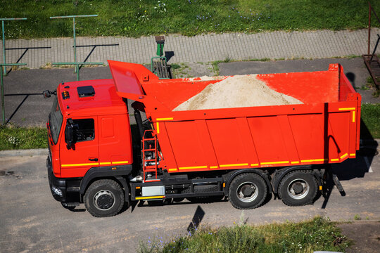 Bright red dump truck with open bed loaded with large pile of sand, parked on asphalt road, high angle view captures heavy construction vehicle beside paved sidewalk and green grass.