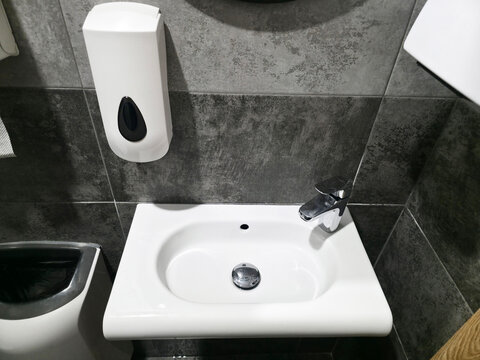 Modern public restroom interior white ceramic sink, chrome faucet, wall mounted soap dispenser against dark grey tiled wall for hygiene handwashing.