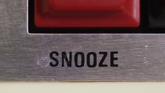 Finger presses a red SNOOZE button on a silver alarm clock for delay and procrastination concepts