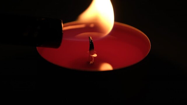 Close-up of red candle being lit with a lighter, burning flame, and glowing wick against a dark black background. High quality FullHD footage