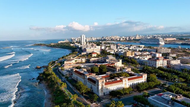 4K pan drone shot of the San Juan coastline near Escuela de Medicina Tropical, Puerto Rico. Shows ocean and cityscape. For tourism, travel, culture.