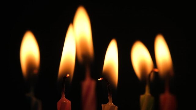 Close-up view of burning birthday candles with bright flickering flames in darkness. Slow motion video.