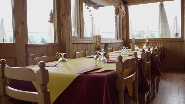 Rustic wooden interior of a cozy alpine lodge dining room in Italy. Set tables by large windows overlooking the mountains inside a traditional mountain chalet or retreat