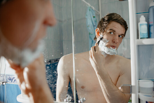 A shirtless man shaves his face with a razor in the bathroom mirror