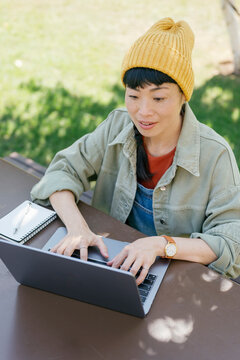 Woman sitting outside using laptop and taking notes in casual outfit