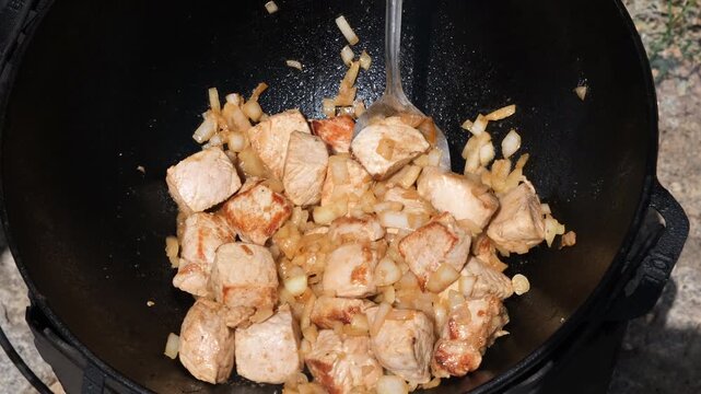 Outdoor cooking of diced pork with chopped onions frying in a traditional cast iron cauldron over an open fire. High quality 4 K footage