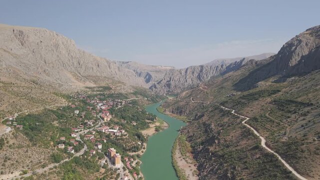 4K 10-bit drone shot flying forward over a riverside village towards the massive stone entrance of the Dark Canyon on a sunny day in Kemaliye, Turkey